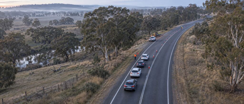 A misty regional highway at dawn, with a traffic controller and a line of cones slowing cars through roadworks.